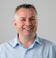 A main with short hair who wears a blue Oxford shirt faces the camera.