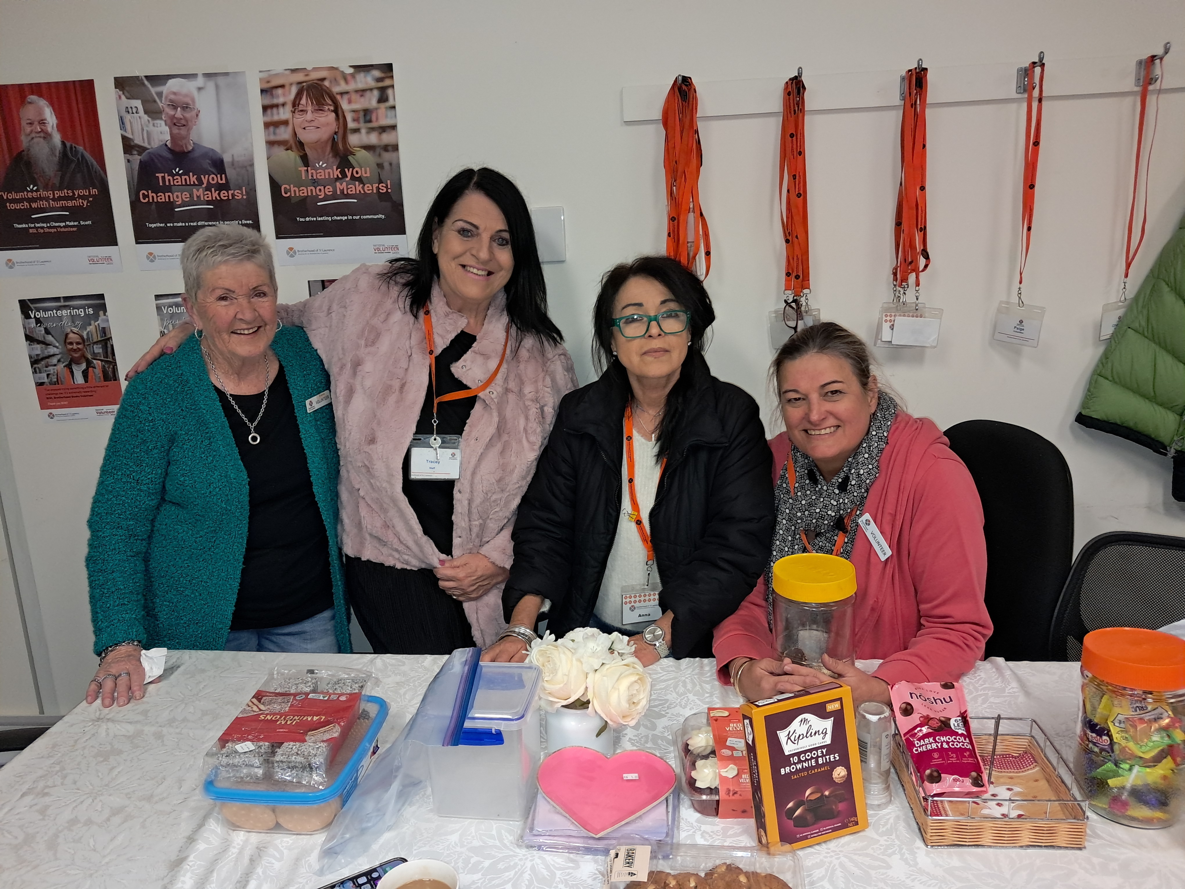 A group of women volunteers from our Belmont op shop are seated in front of a table with gifts, flowers and morning tea, smiling at the camera.