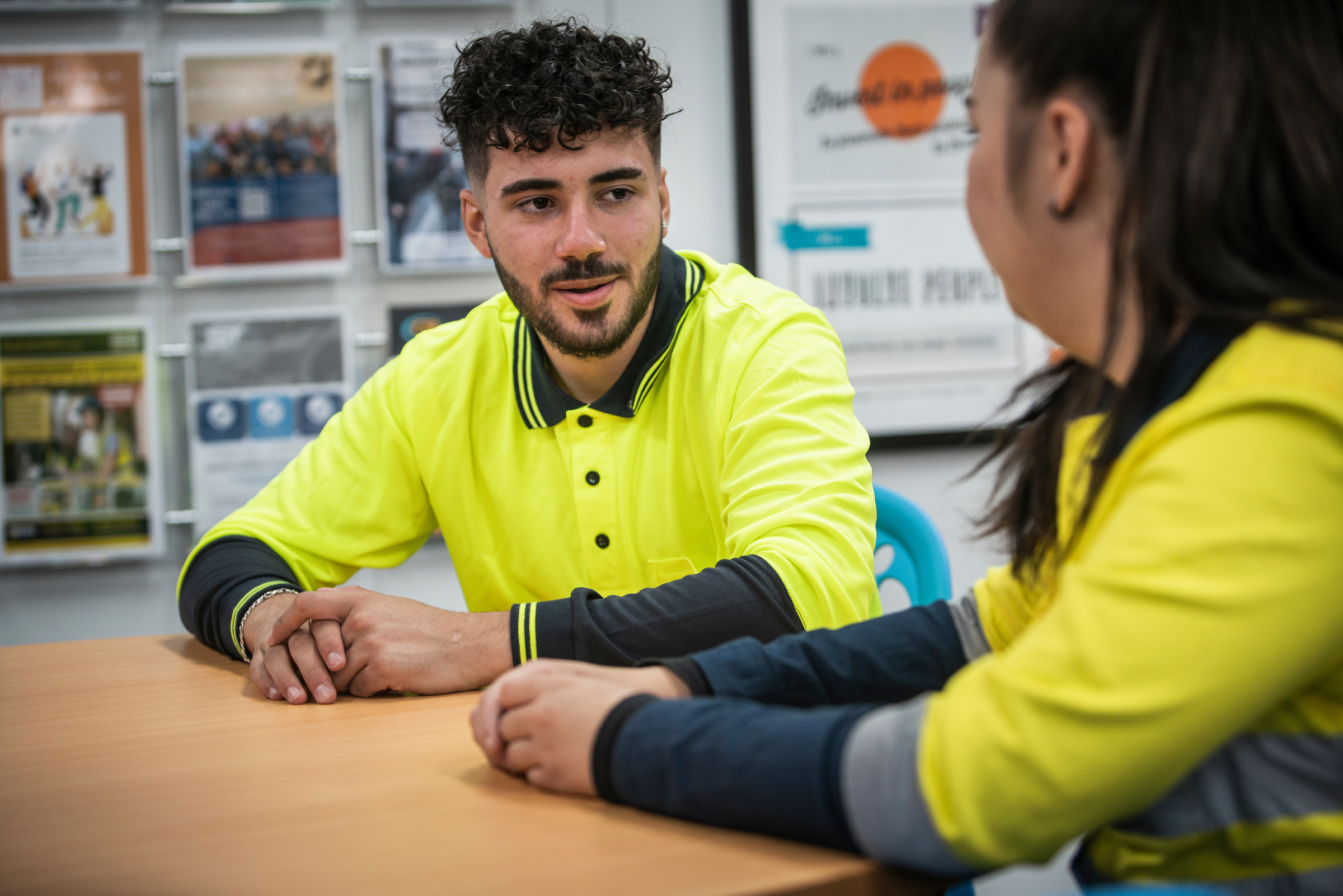 A young apprentice sits at a table having a work conversation with another employee. Both are dressed in Hi-Vis clothing.