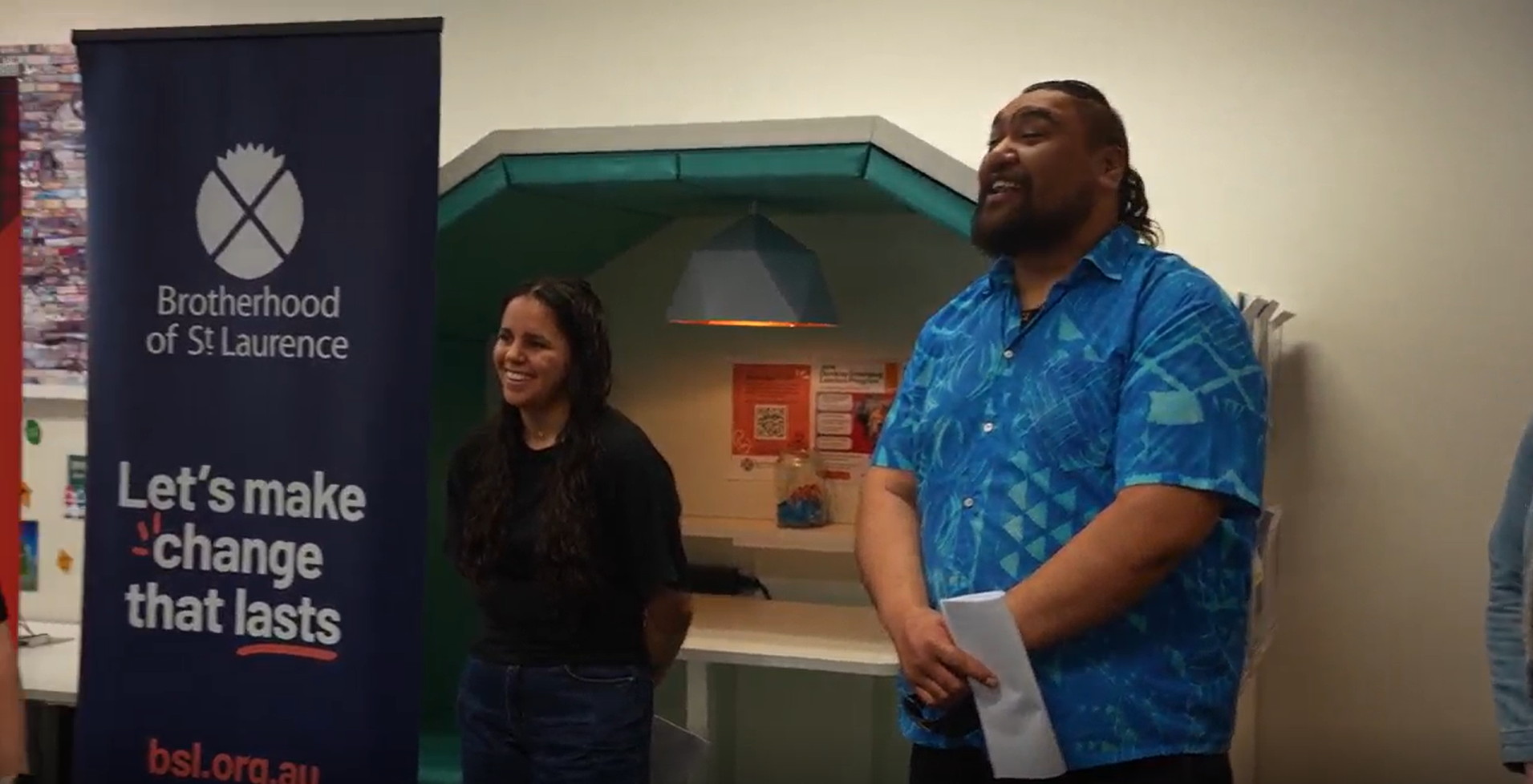 Obeda's story - Two people standing indoors near a Brotherhood of St. Laurence banner, one holding notes, during a presentation moment.