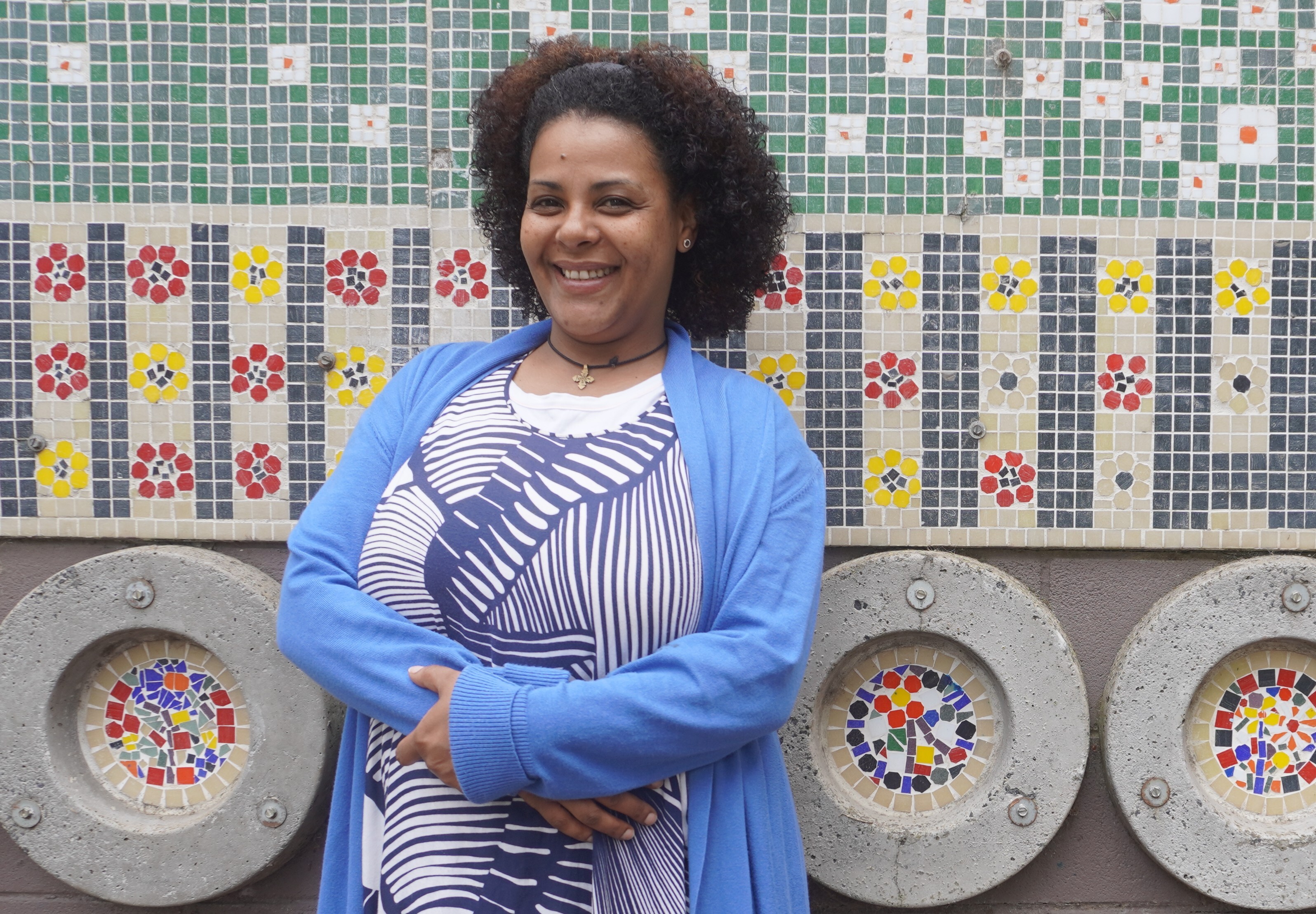 Beza's story - Woman standing with arms crossed in front of a colourful tiled wall at a community centre, wearing a patterned top and blue cardigan.