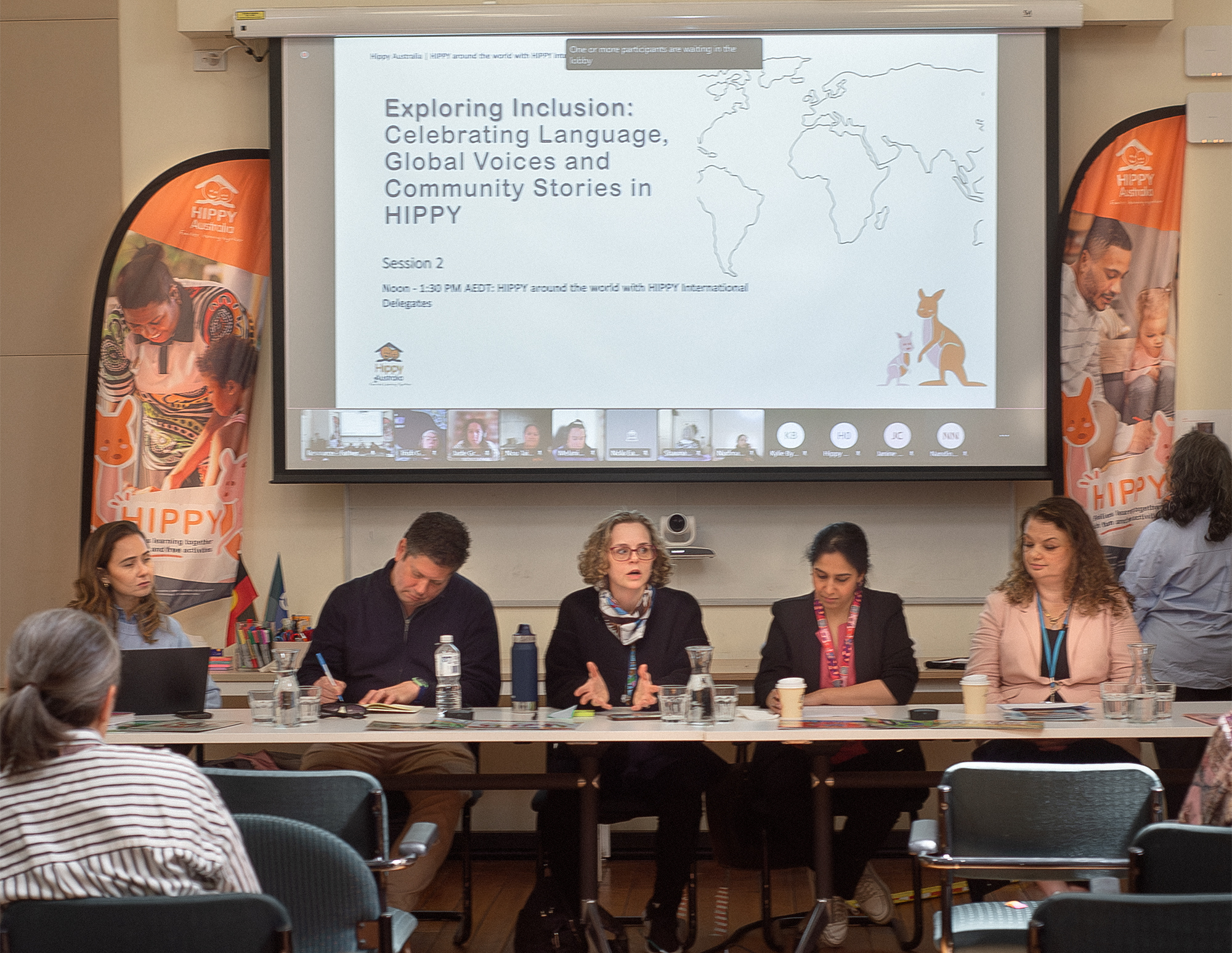 Five HIPPY panellists sit at a long table during a HIPPY conference session on inclusion, with a presentation screen and banners behind them.