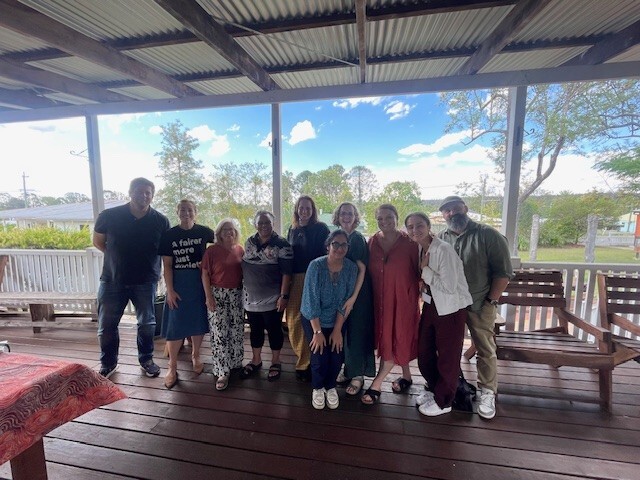 A group of ten people standing together on a covered wooden deck, posing for a photo with trees and a bright blue sky in the background.