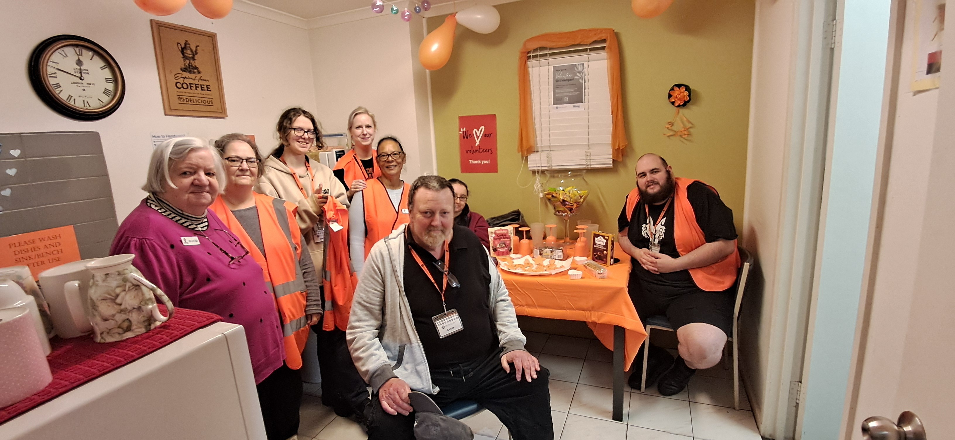 A group of male and female volunteers from our Hoppers Crossing op shop are smiling at the camera while seated at a table with gifts and morning tea.