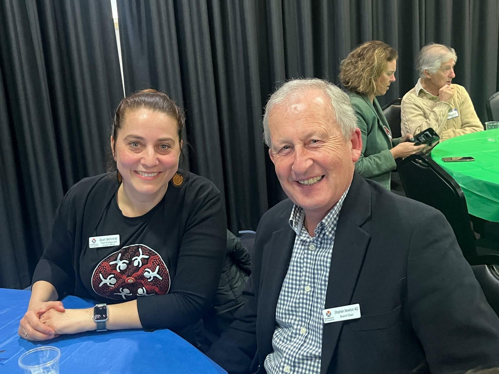 Left to right: BSL Chief Strategy and Impact Officer Zeah Behrend and BSL Board Chair Stephen Newton are seated at a table smiling for the photo. Zeah has brown hair and is wearing a black t-shirt with an indigenous design on it. Stephen is wearing a navy check shirt and blazer.