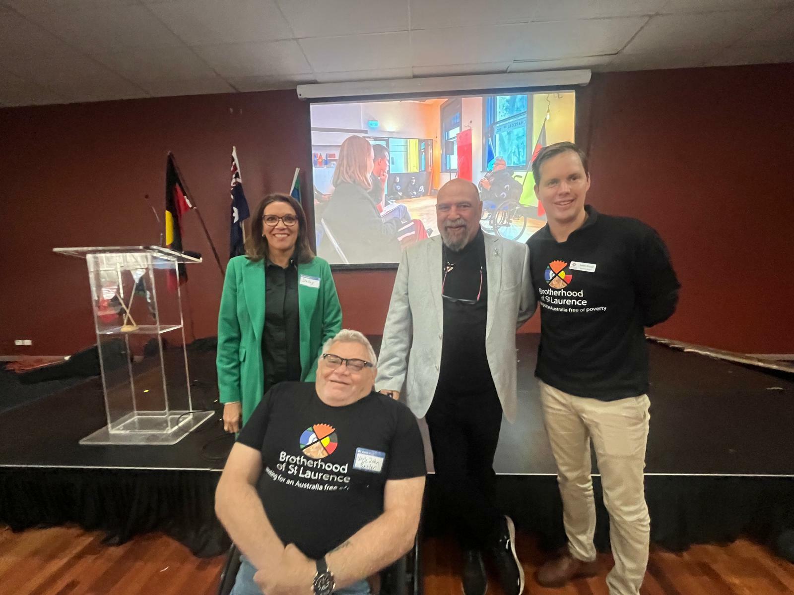 Left to right: MC Shelley Ware, guest speaker Richard Frankland, BSL Cultural Ambassador Uncle John Baxter, and BSL Executive Director Travers McLeod are in front of the stage, with a TV screen behind them which has a photo featuring a BSL event. Shelley has brown hair and is wearing a green jacket; Richard has a beard and is wearing a camel jacket and Uncle John and Travers are wearing BSL t-shirts. They are all smiling at the camera.