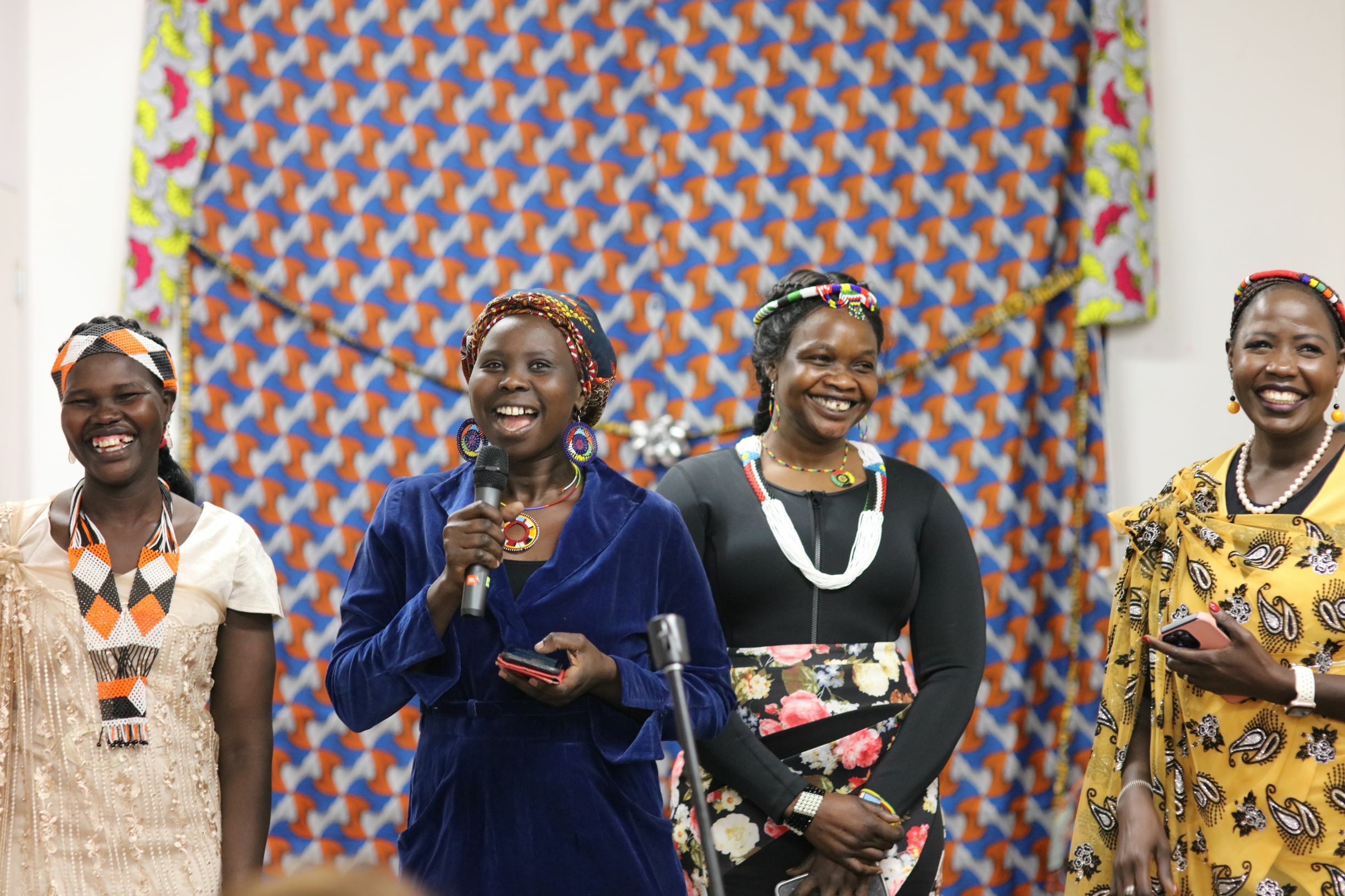 Women of the Well from left: Mary Deng, Nyibol Deng, Martha Chol and Mary Top They are standing in front of a colourful curtain and smiling.