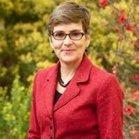 A photograph of Jenny Trethewey. She is standing infront of trees and she is wearing a red jacket.