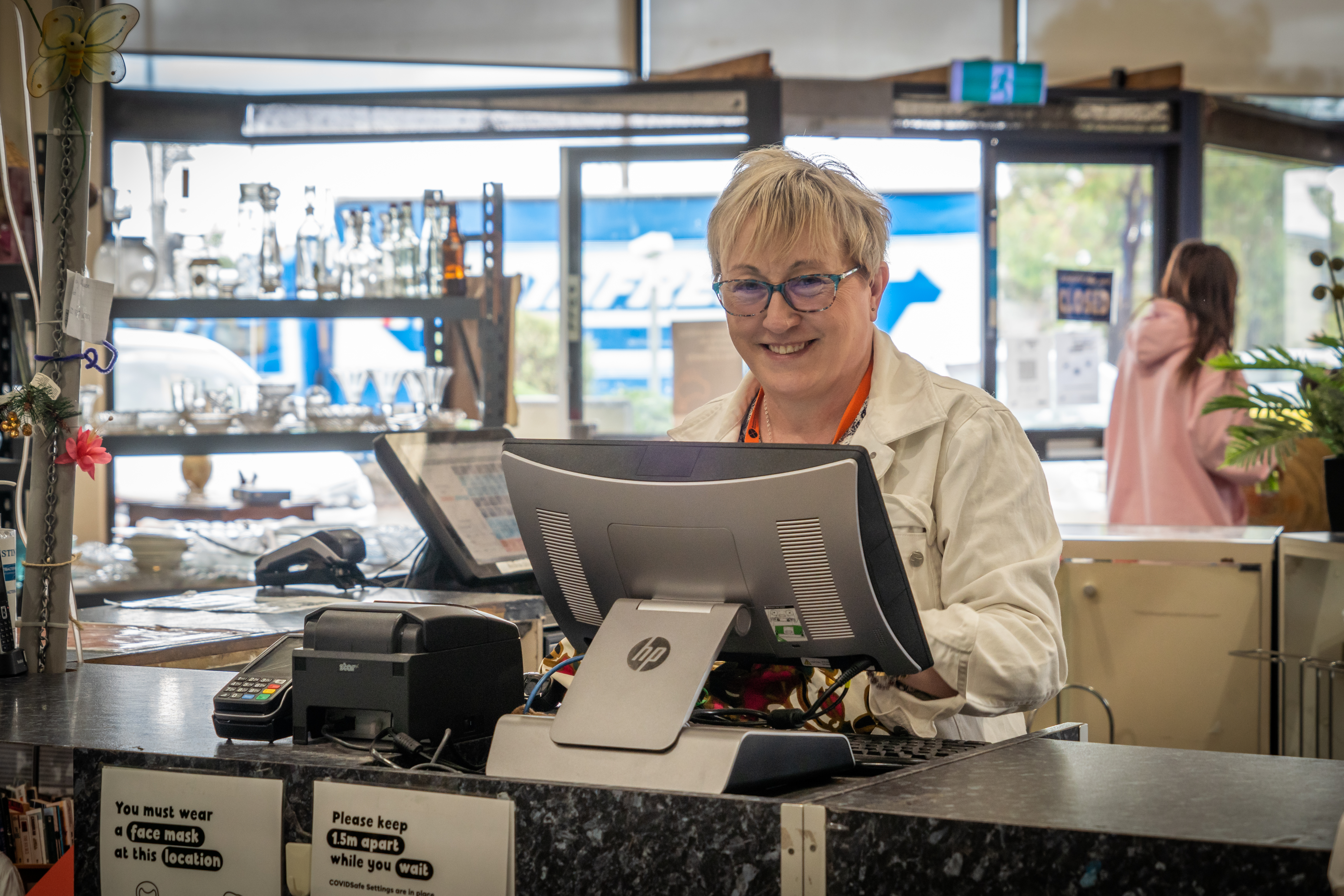 A woman with blonde hair behind a BSL Op Shop cashier counter smiling