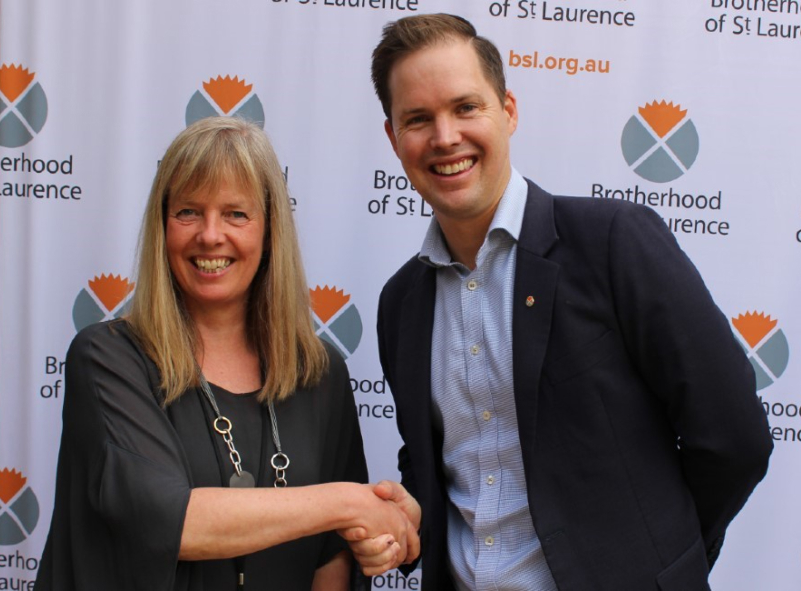 A photo of a woman with long blond hair and a man shaking hands wearing a suit. They are standing in front of a banner which has the BSL log on it and smiling.