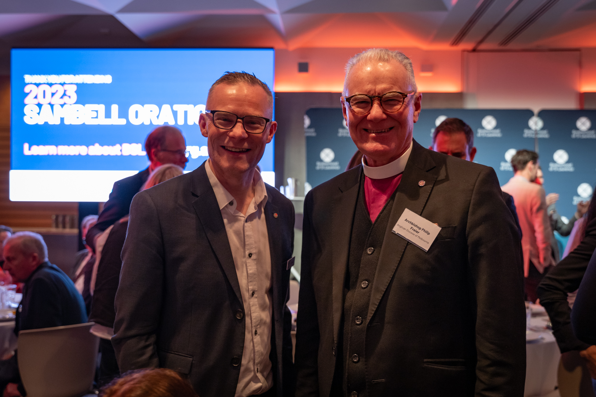 BSL’s Internal Communications Lead David Bain, and The Most Reverend Dr Philip L. Freier. They are smiling at the camera and behind them is a digital screen with the words 2023 Sambell Oration on it