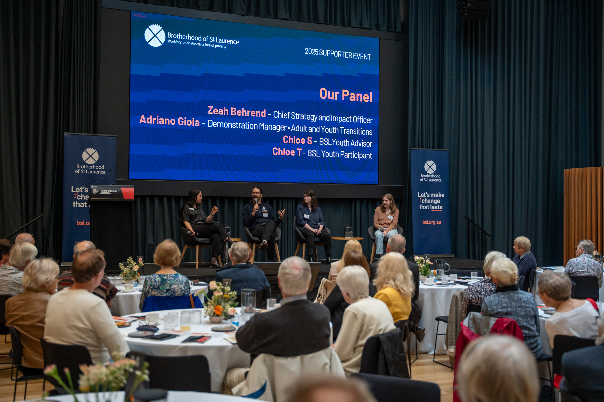 Four people discuss a panel subject on stage in front of tables of people sitting down and watching and listening.