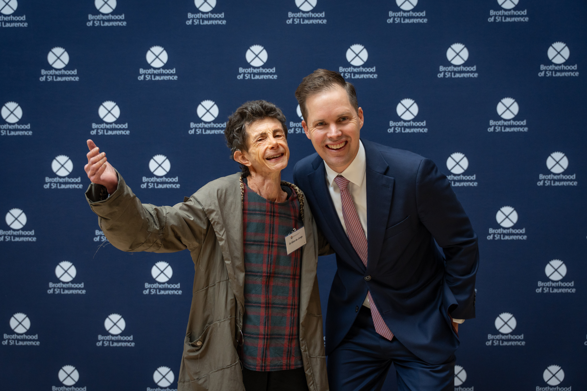 Two people smiling and gesturing in front of a media wall with Brotherhood of St. Laurence branding.