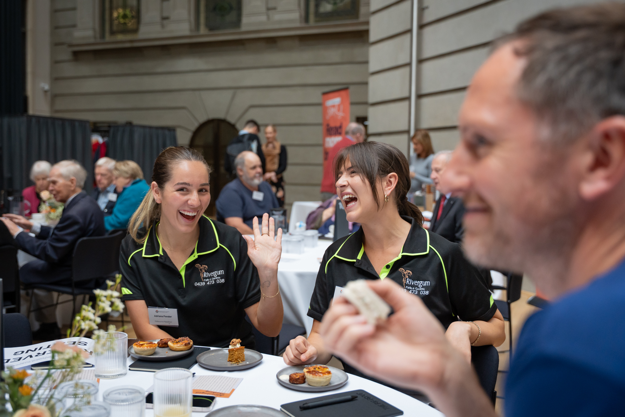 Two female-presenting people laugh and smile over quiche while sitting at an event table.