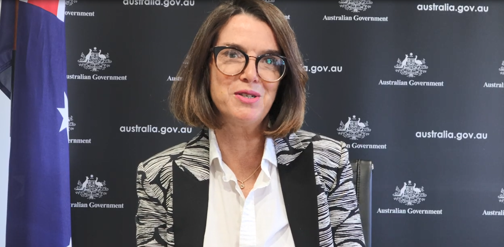 A woman sits in front of an Australian Government screen and beside a single Australian flag