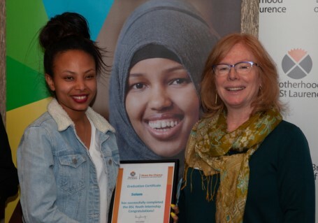 This is a photo of Selam receiving her Internship graduation certificate from Conny Lenneberg, the Executive Director at Brotherhood of St. Laurence. The photo was taken in 2019. Selam and conny are standing in front of pictures of participants and a backdrop showing the Brotherhood of St. Laurence logo.