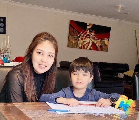 This is a photo of Zahra and her son. They are both smiling and sitting at the dining table. Zahra is helping her son with some remote learning tasks.