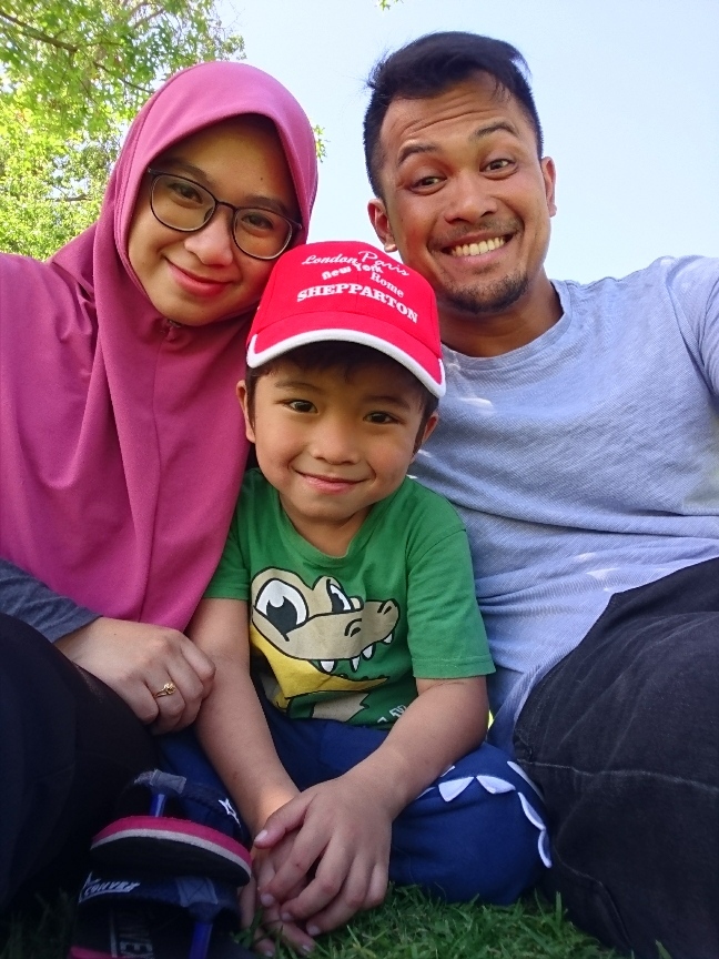 This is a photo of Ammar and his family, including his wife and young son. They are smiling and sitting on grass. There is a tree in the background.