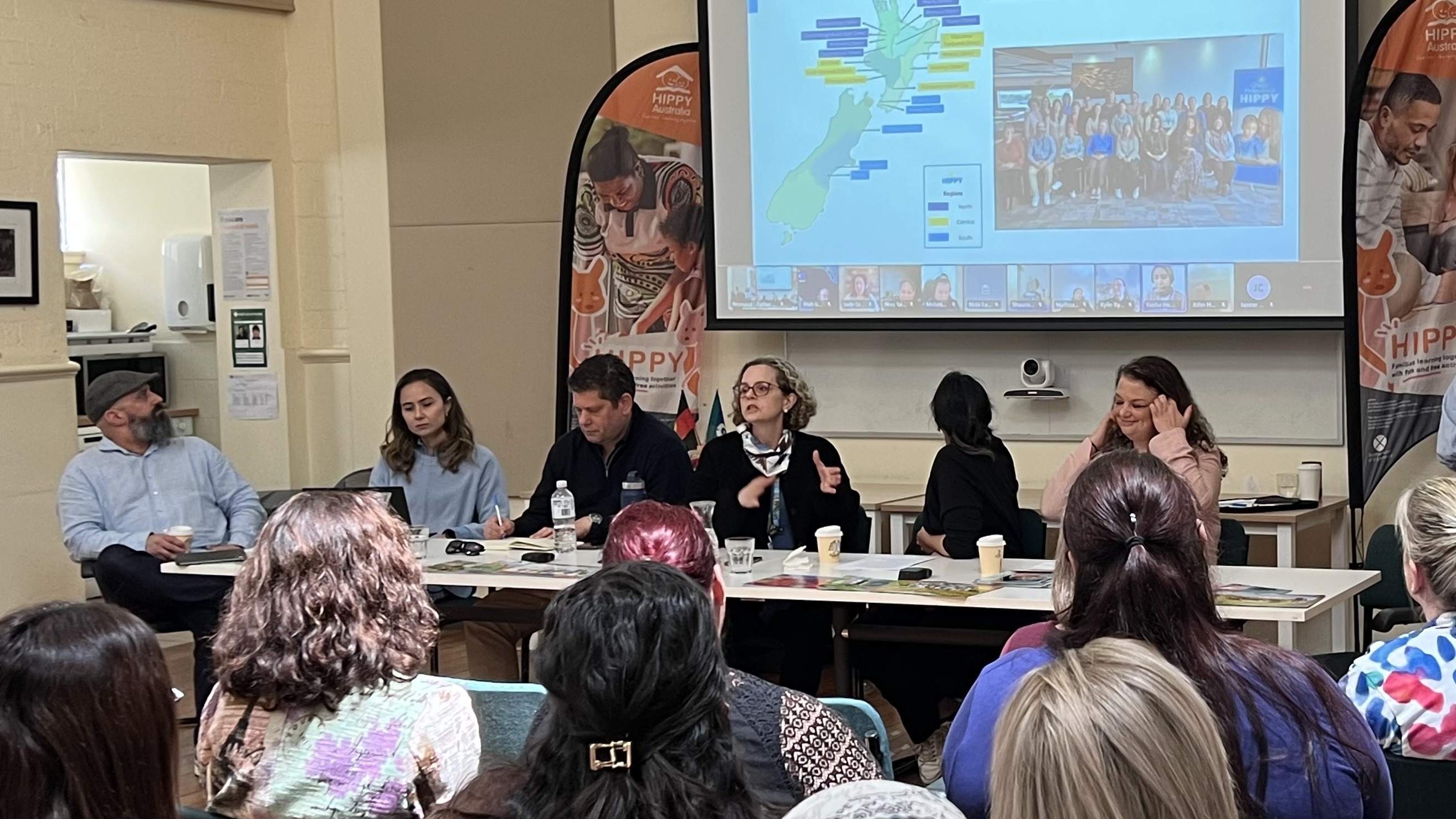 A panel discussion in a community setting with six HIPPY panellists seated at a long table, facing an audience, and a presentation projected on a screen behind them.
