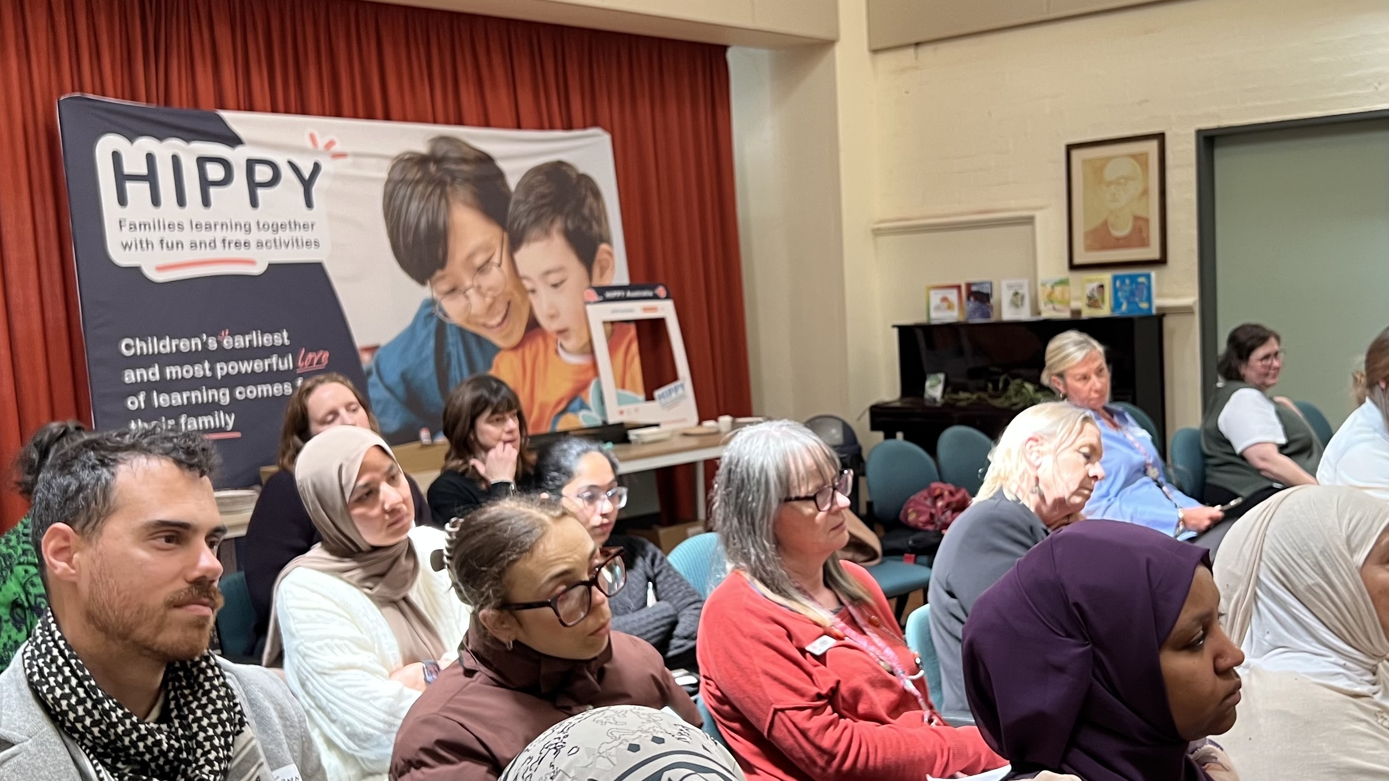 Adults sit attentively in a community room during a HIPPY presentation, with a banner and educational materials displayed behind them.