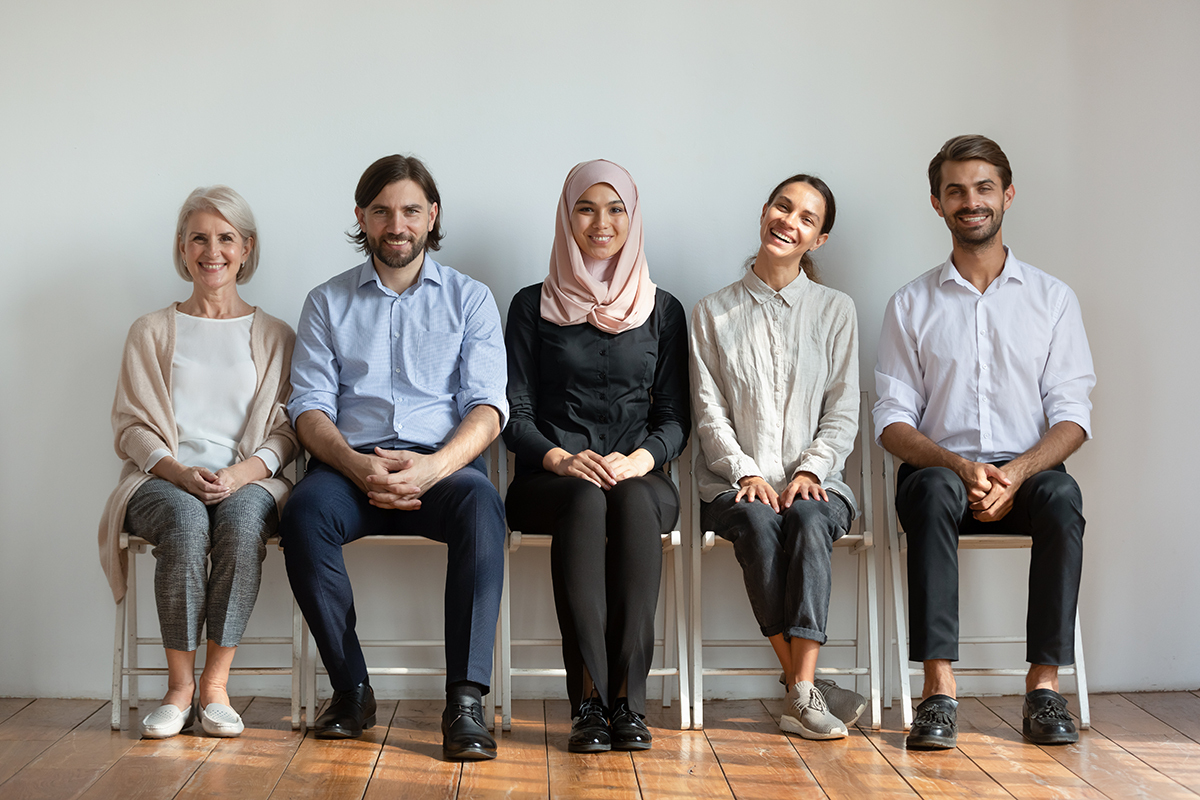 Smiling multicultural young and old professional business people sit on chairs in row looking at camera, happy staff job candidates group team portrait, human resource, diverse ethnicity concep
