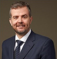 A man with short brain hair and a beard faces the camera while wearing a suit and tie.